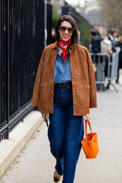 Guest wears brown suede jacket, navy blue denim jeans, orange bag, blouse, red scarf outside Lacoste during the Womenswear Fall/Winter 2025/2026 as...