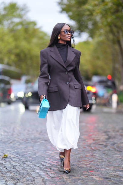 Guest wears brown sunglasses, a black turtleneck pullover, black blazer jacket, a white skirt, a light blue leather Balenciaga bag with crocodile...
