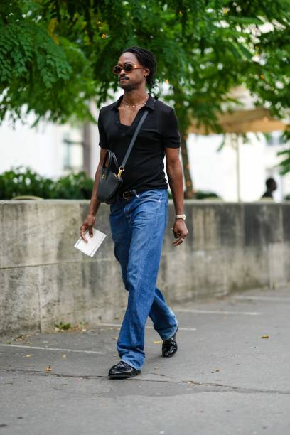 Guest wears brown sunglasses from Celine, a gold necklace, a black short sleeves t-shirt, a black matte leather crossbody bag, blue denim large...