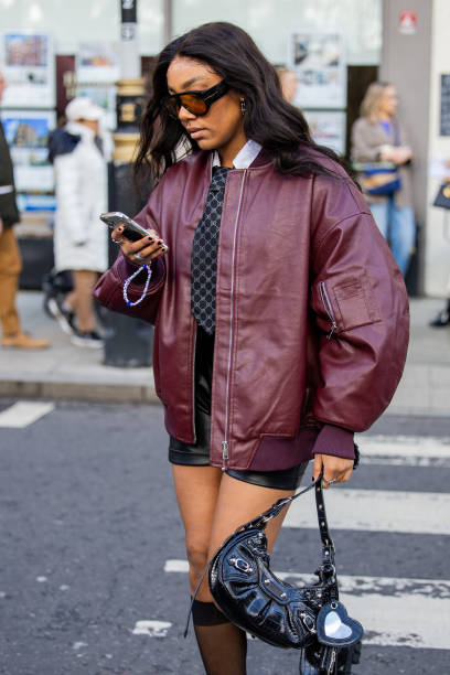 Guest wears burgundy dark red bomber jacket oversized, black Balenciaga bag, tie, shorts outside Bora Aksu during London Fashion Week February 2024...