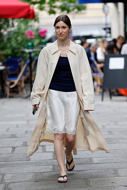 Guest wears dark blue top, white silk skirt with lace, beige trench coat, outside Lemaire, during the Menswear Spring/Summer 2026 show as part of...
