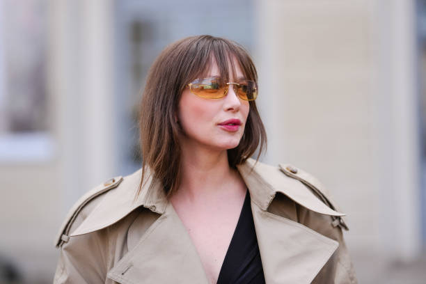 Guest wears dark orange sunglasses, black deep v-neck shirt, light brown trench coat, outside Tod’s, during the Milan Fashion week Women's...
