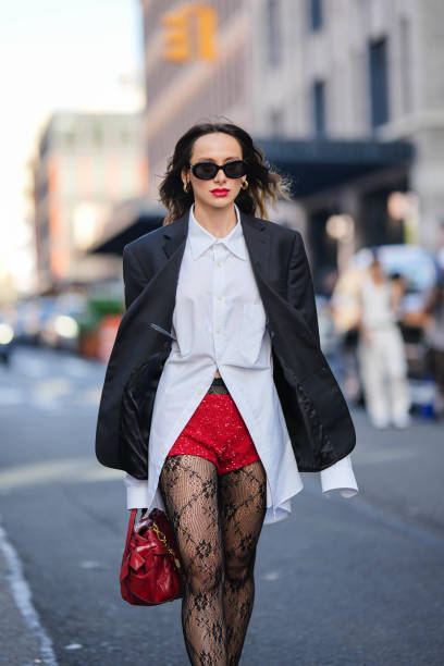 Guest wears earrings, sunglasses, a white shirt, a black oversized blazer jacket, red sequined mini shorts, black mesh tights with floral embroidery,...