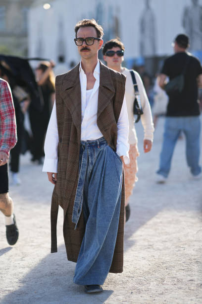 Guest wears glasses, a brown sleeveless long checked trench coat, a white shirt, wide leg blue denim jeans, outside Casablanca, during the Womenswear...