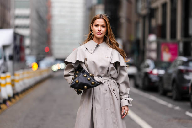 Guest wears gold earrings, light gray Altuzarra belted trench coat, shiny black large bronzed studs design Altuzarra origami leather bag, outside...