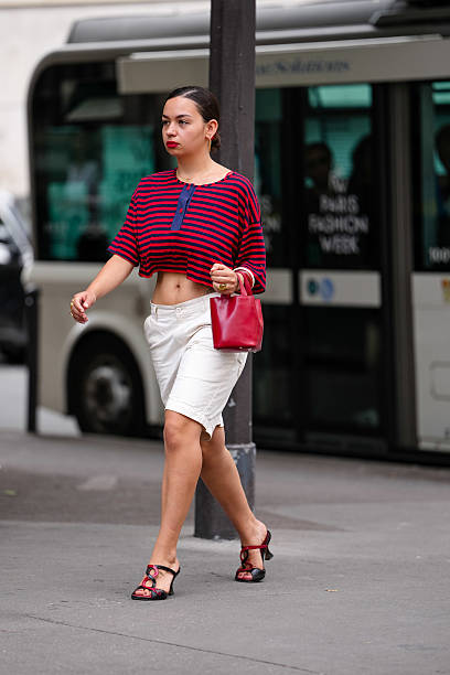 Guest wears gold hoop earrings, bright cherry red black striped short sleeve cropped shirt, gold rings, shiny red leather bag, white shorts, black...