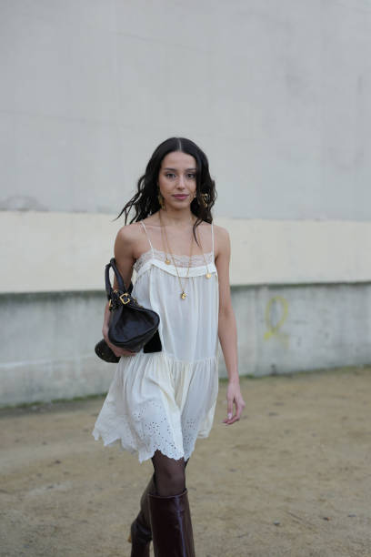 Guest wears gold necklace, white lace Chloe mini dress, shiny black Chloe leather bag, black sheer mesh tights, outside Chloe, during the Paris...