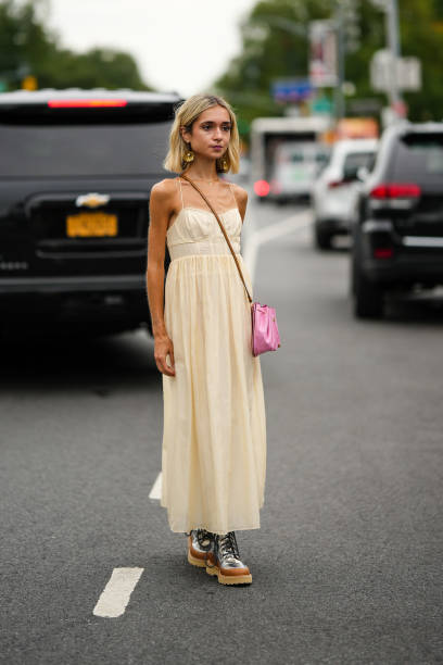 Guest wears gold pendant earrings, a beige square neck long dress, a pale pink shiny leather crossbody bag, silver shiny leather laces ankle boots,...