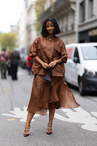 Guest wears gold pumps embellished with rhinestones, a brown leather mini bag, a gold necklace and a brown coord set outside Akris show during...