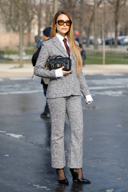 Guest wears grey printed Chanel suit, white striped shirt, burgundy tie, black Chanel bag, black heels, outside Chanel, during the Womenswear...