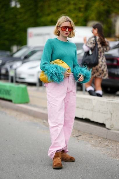 Guest wears orange aviator sunglasses, a gold necklace, a green braided wool with embroidered feathers wrists pullover, a yellow shiny braided...
