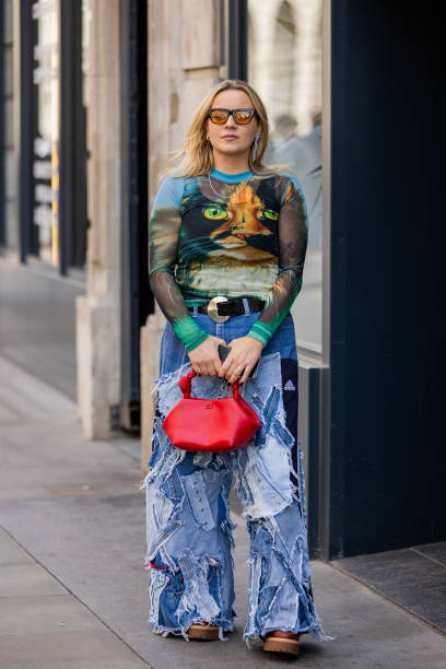 Guest wears patched denim jeans, red bag, shirt with graphic print outside Bora Aksu during London Fashion Week September 2024 on September 13, 2024...