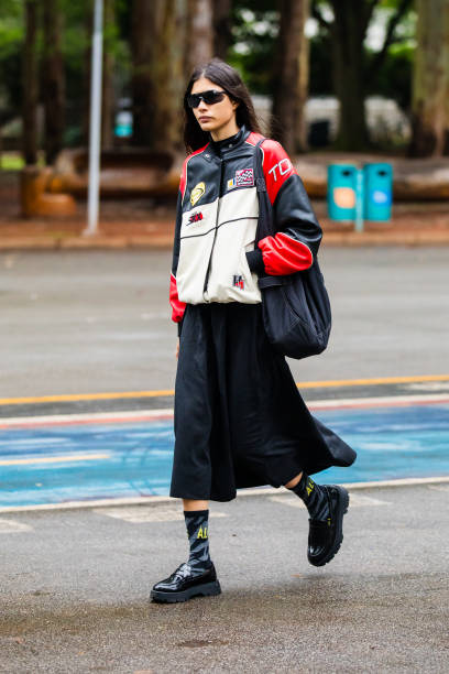 Guest wears racing jacket, black midi skirt, black chunky shoes, black socks with yellow lettering, black oversized bag, black sunglasses at Sao...