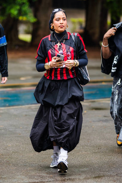Guest wears red and black football jersey, black pleated skirt, gold jewelry, white socks, chunky sneakers at Sao Paulo Fashion Week 2024 N58 -...