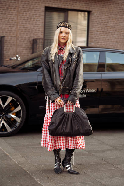 Guest wears red and white checkered dress, black and white pointy shoes, black bag, brown leather jacket, knitted hat outside A. Roege Hove show...