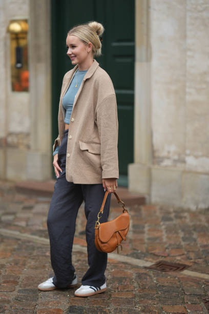 Guest wears silver earrings, a blue gray cropped t-shirt, a beige blazer jacket, gray suit pants, white sneakers, a camel shiny leather Saddle...