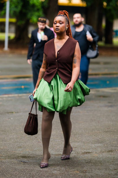 Guest wears sleeveless brown vest, green balloon skirt, beige tights, purple shoes, brown handbag, braided orange hair at Sao Paulo Fashion Week 2024...