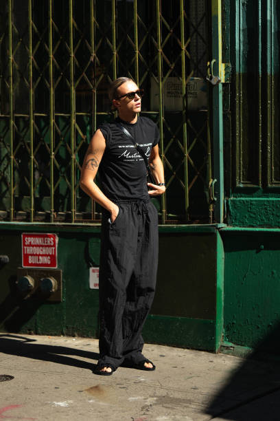 Guest wears sunglasses, a black t-shirt worn as a tank top, black slack palazzo pants, sandals, outside Brandon Maxwell, during New York Fashion...