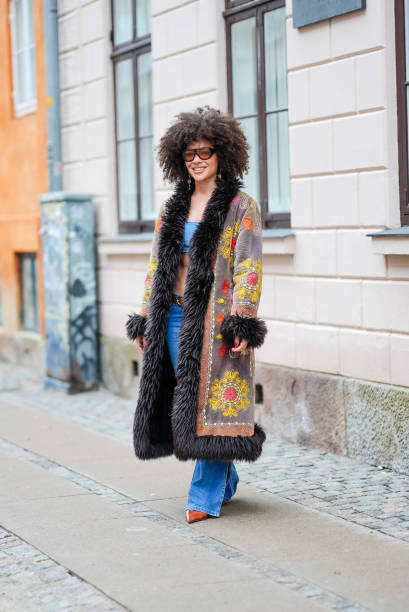 Guest wears sunglasses, a blue bras, a gray and black long winter coat with fluffy parts and printed geometric patterns in yellow and orange, a belt,...