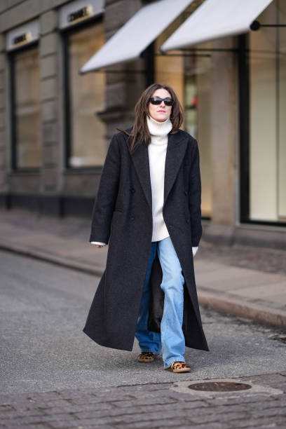 Guest wears sunglasses, a white oversized turtleneck ribbed wool pullover , a black oversized long coat , blue flared denim jeans pants , leopard...