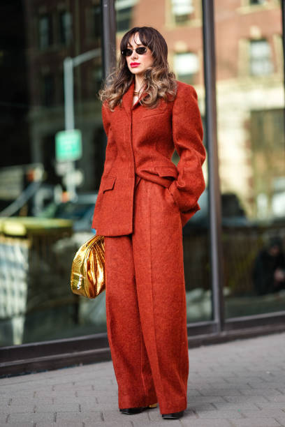 Guest wears sunglasses, an orange oversized blazer jacket with shoulder pads, cotton pants , a golden shiny bag, outside Carolina Herrera, during New...