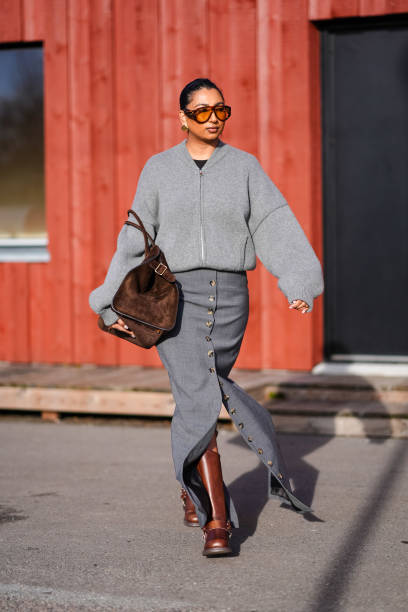 Guest wears sunglasses, earrings, a gray wool oversized cardigan, a brown suede bag, a gray long slit skirt , brown leather boots , outside Gestuz,...