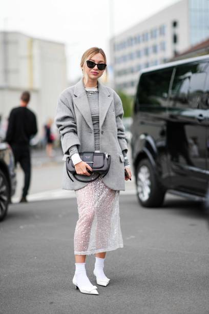 Guest wears sunglasses, necklaces, a grey pullover, a grey fluffy jacket, a two-tone Prada bag, a sequined white transparent lace midi skirt, white...