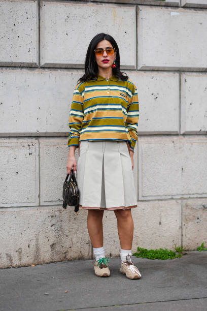 Guest wears sunglasses, red lp, a khaki and yellow striped polo shirt, a knee length pleated skirt, a bb leather bag, socks, sneakers shoes, outside...