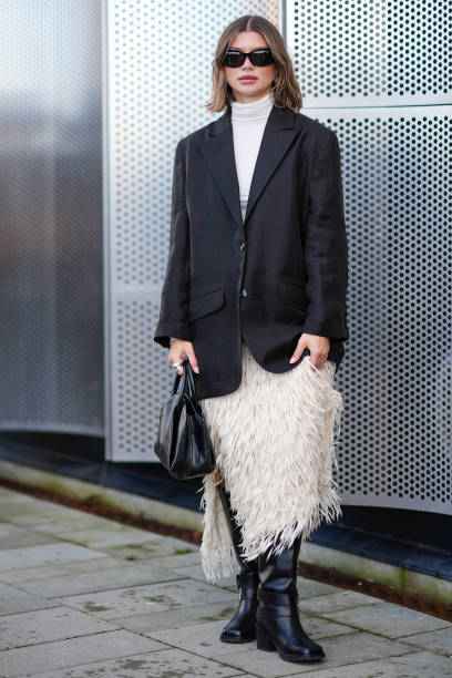 Guest wears sunglasses, silver earrings, a piercing, a white turtleneck, a black oversized blazer jacket, a white fluffy fringed skirt , a black...