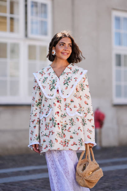Guest wears white earrings, cream and white floral pattern/print oversize long sleeve shirt, light purple midi skirt, light brown bag, silver rings,...