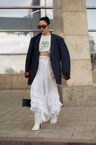 Guest wears white long maxi skirt, white Adidas socks, cropped beige top with green letters and navy blue oversized blazer on day 1 of Visa Fashion...