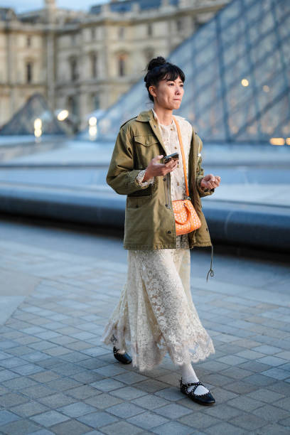 Guest wears white sheer mesh lace midi dress, bright orange bag, dark green oversized jacket, white socks, black ballerina flats leather shoes,...