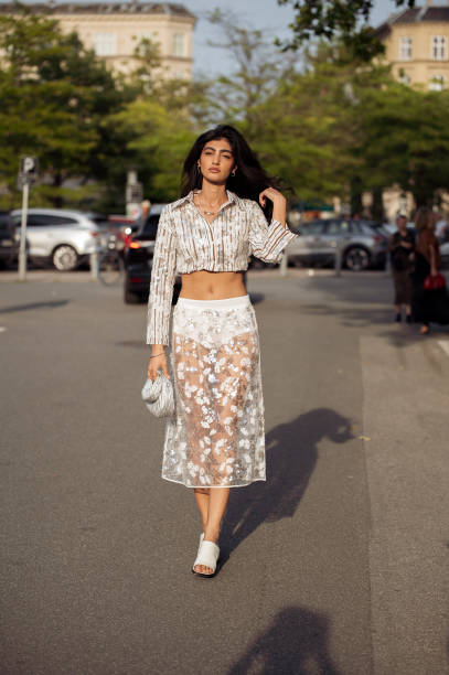 Guest wears white sheer skirt with flowers and sparkles, matching crop long sleeve top, two silver Miu Miu bags and white shoes outside the Baum und...