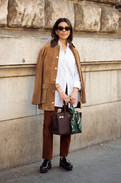 Guest wears white shirt, brown pants, brown suede jacket and brown Hermes bag with a green and white silk neck scarf outside the Hermes fashion show...