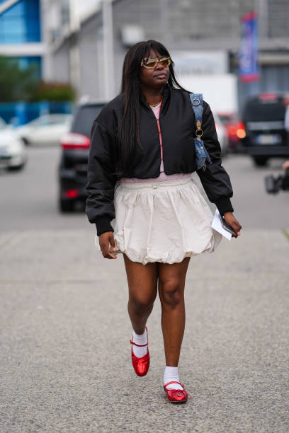 Guest, wears yellow lined glasses with dark brown lens, black cropped bomber jacket, pink shirt, white skirt, white socks, red ballet shoes,outside...