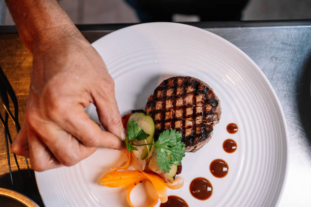 hand of a chef garnishing a fillet of beef with herbs and vegetables on a ceramic plate in a kitchen - food stock pictures, royalty-free photos & images