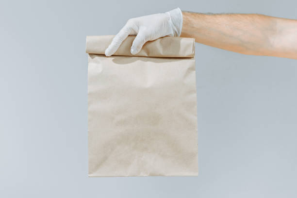 hand of a man in a glove holding a brown bag or bag for taking out isolated on a gray background. packaging layout template. delivery service concept. copy space. courier - junk food stock pictures, royalty-free photos & imag