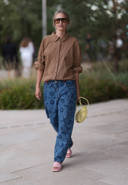 Hanni Gohr is seen wearing angular aviator sunglasses with a black frame and orange lenses, white dangling chain earrings, a buttoned brown and beige...