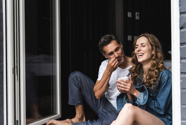 happy couple in nightwear at home sitting at french window - coffee stockfoto's en -beelden