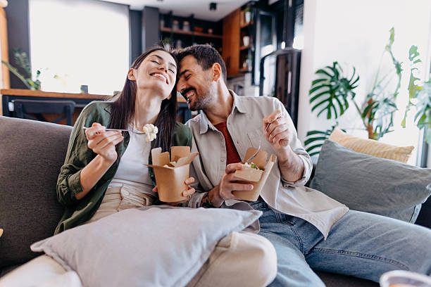 happy couple laughing and eating takeaway food at home - junk food stock pictures, royalty-free photos & images