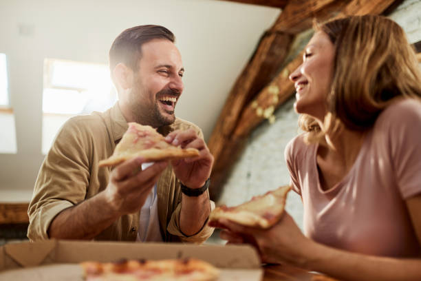 happy couple talking while eating pizza for lunch at home. - junk food stock pictures, royalty-free photos & images