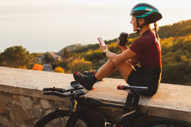 happy cyclist woman relaxing after long ride, drinking coffee, smiling and chatting in mobile smartphone at sunset - junk food stock pictures, royalty-free photos & images