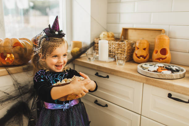 happy laughing baby girl with bowl of popcorn on kitchen, decorated for halloween - home decoration stock pictures, royalty-free photos & images