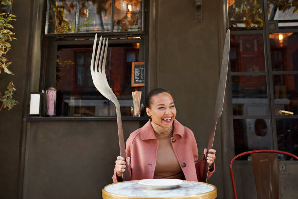 happy woman holding large fork and table knife while sitting at sidewalk cafe - food stock pictures, royalty-free photos & images
