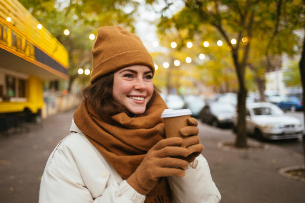 happy woman wearing knit hat day dreaming with disposable coffee cup on street during winter - junk food stock pictures, royalty-free photos & images