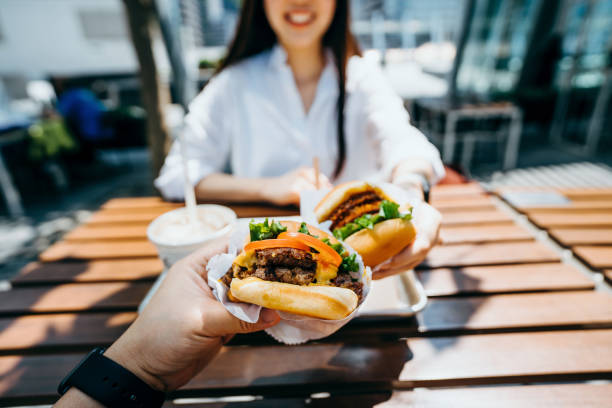 happy young asian couple eating beef burger with fries in an outdoor fast food restaurant - junk food stock pictures, royalty-free photos & images