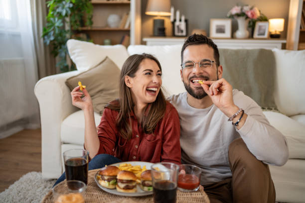 happy young couple eating fast food at home and watching movie on tv - junk food stock pictures, royalty-free photos & images