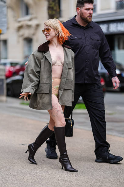 Hayley Williams wears orange sunglasses, dark green oversized jacket with dark brown collar, cream bralette, cream mini shorts with floral detail,...