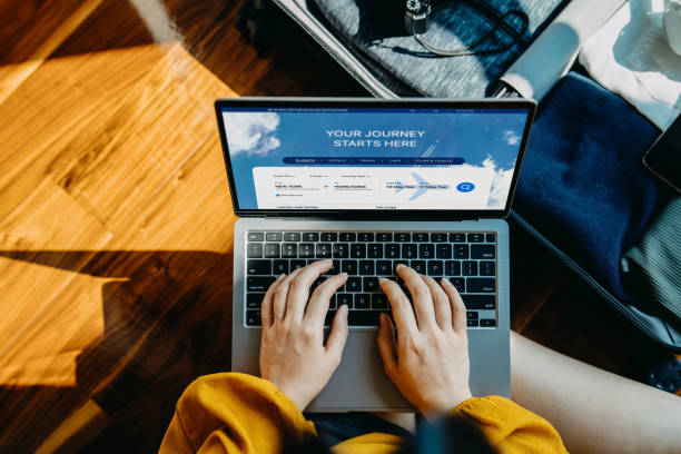 high angle shot of young asian woman booking flight tickets on airline website online with laptop, with packed suitcase by her side. travel planning. booking a holiday online. travel and vacations concept. ready to travel. bu
