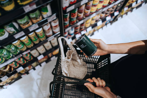high angle view, close up hands of young asian woman grocery shopping in supermarket. she is putting a tin can into a cotton mesh eco bag in a shopping basket. environmentally friendly and zero waste concept - food fotografía